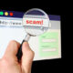Forgery and Fraud - A hand holds a magnifying glass over the location bar of a browser, revealing the URL is a "scam".